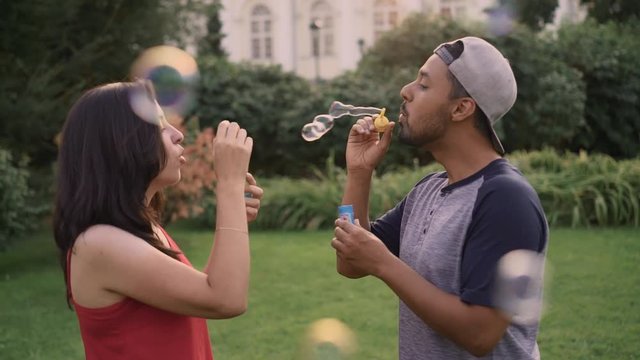 Beautiful young couple blowing soap bubbles at each other on sunny summer day. Concept of romance and fun. Handheld slow motion medium shot