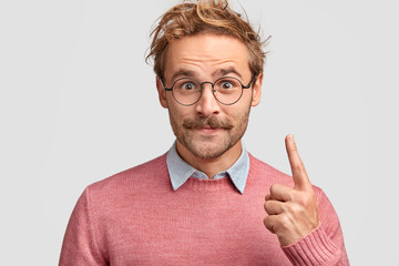 Horizontal shot of handsome satisfied hipster has mustache, dressed in elegant clothes, points with...