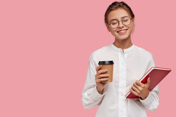 Dreamy pleased Caucasian woman with joyful expression, has charming smile on face, drinks takeaway coffee and notepad, stands against pink background with free space for your advertisement or text