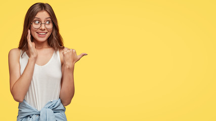 Charming happy Caucasian girl with dark hair, smiles broadly, indicates with thumb aside, suggests great place for friend, has shirt around waist, isolated over yellow background. Promotion. - obrazy, fototapety, plakaty