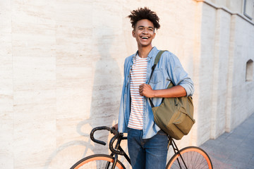 Happy young african guy walking with bicycle outdoors on the street.