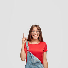 Indoor shot of good looking satisfied brunette woman has pleased facial expression, gets brilliant idea in mind, points with fore finger upwards, dressed casually, isolated over white background