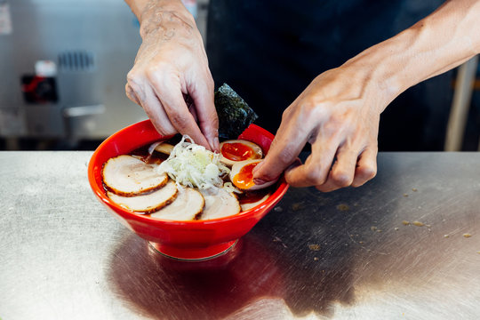 Chef plating Shoyu Chashu Ramen: Japanese ramen Shoyu sauce soup with chashu pork, boiled egg, dry seaweed and chives in red bowl