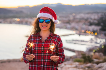 Fototapeta na wymiar Holiday, Christmas and people concept - Young happy woman in Santa hat over beautiful landscape with fireworks and sparklers