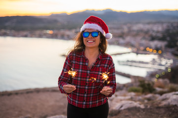 Christmas, holidays, people concept - woman in Santa hat and sunglasses holding sparklers over sea background