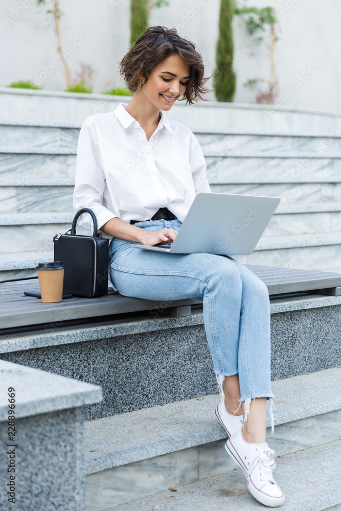 Wall mural Young beautiful woman sitting outdoors using laptop computer.