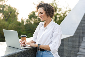 Obraz premium Cute beautiful woman walking outdoors using laptop computer holding coffee.