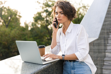 Fototapeta premium Displeased beautiful woman standing outdoors using laptop computer talking by phone.