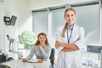 Obraz premium I love my job. Waist up portrait of beautiful lady in white lab coat posing in gynecological cabinet while red-haired woman looking at her with smile