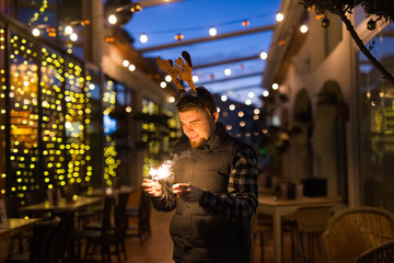 Fototapeta premium Christmas and holiday concept - Happy man in Christmas deer costume with sparkler
