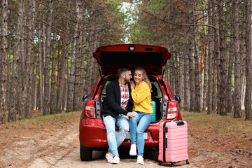 Young couple sitting in car trunk and suitcase on forest road