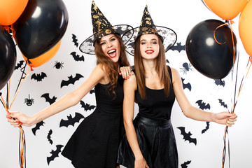 Halloween party. Two gorgeous women in black dresses and witch`s hats hold black and orange balloons on the background of the wall with bats.