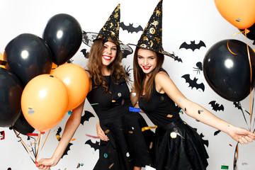 Two beautiful girls in black dresses and witch`s hats have fun with black and orange balloons and confetti. Halloween party