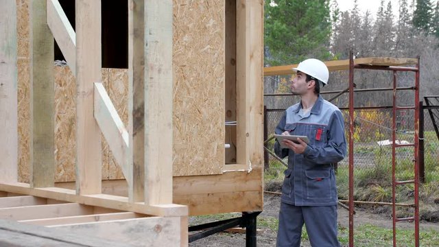 Caucasian worker in front of a house. Architect or builder check plans in a half built timber frame house. Builder on a construction site with a tablet. 4k