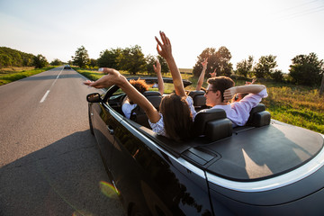Black cabriolet is on the country road. Company of young girls and guys are sitting in the car hold their hands up on a sunny day.