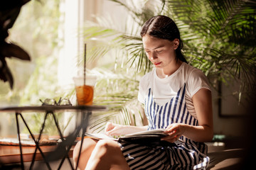 A young slim friendly girl with dark hair,dressed in casual outfit,sits at the table and reads a book in a cozy coffee shop.