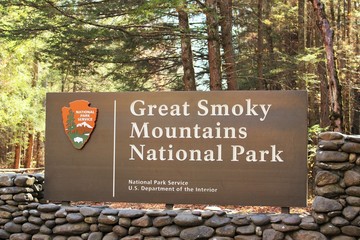 Daytime photo of a Great Smoky Mountains National Park entrance sign with stone surrounding and trees and sunshine in the background