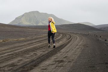 Young woman in yellow jacket running on the black lava field on the road to Landmannalaugar area, Iceland