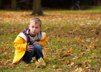 Young boy in yellow jacket crouching over an acorn branch in the park in the autumn, while looking...