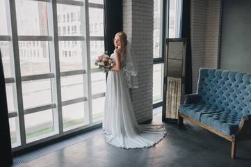 A beautiful bride is standing near a window in a gray studio with a gorgeous bouquet. Wedding portrait. Wedding interior. Wedding photography.