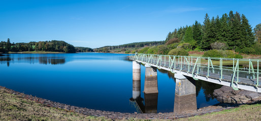 Panorama of Kennick Reservoir in Dartmoor National Park, England, UK, on a bright clear day with the intake tower gantry stretching out, reflections in the water and trees and boats in the background.