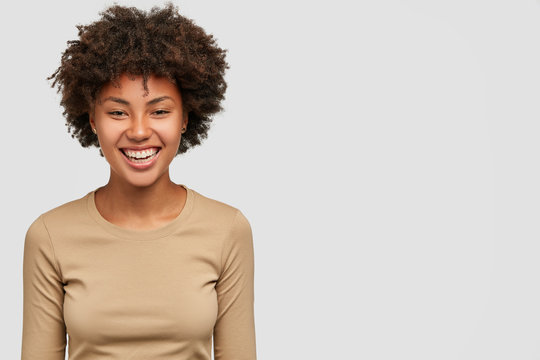 Happy cheerful dark skinned girl smiles gently, satisfied with result of exam she passed, dressed in casual beige sweater, isolated over white background with free space on right side for promotion
