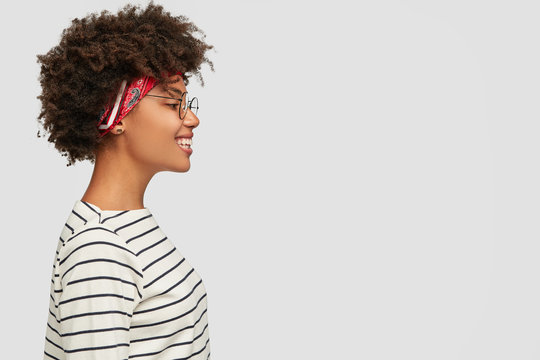 Profile of happy dark skinned girl wears striped clothes, headband, spectacles, smiles positively, isolated over white background for advertisement, promotion or slogan. Black woman stands sideways