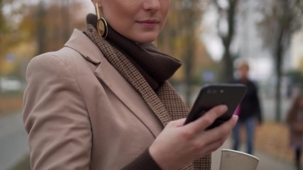 stylish woman typing message and drinking coffee in autumn street