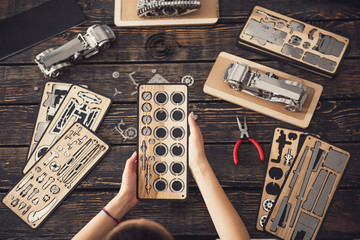 Curious young woman sitting at the table and making model cars with the help of a constructor and pliers