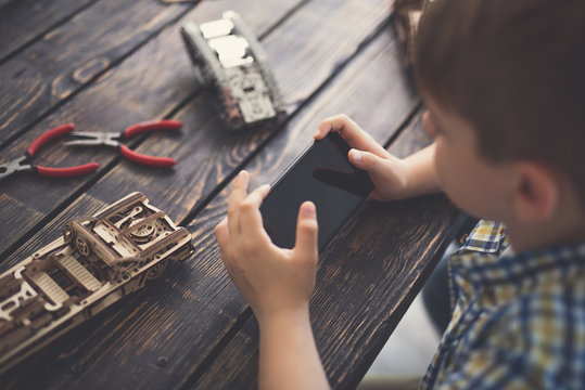 Boy with smartphone. Clever progressive boy sitting alone with wooden constructor on the table and holding modern gadget in his hands