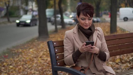 young female uses smartphone while sitting on bench against background of city