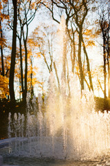 Splashing fountain in autumn park, sunset on background, close up. Beautiful sunset through water fountain, vertical. Autumn time in the city park