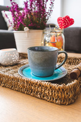 Close up view of a blue vintage cup of coffee on a plate, a candle, apples in a jar with light chain, heart shaped lollipops and calluna flowers in a room. Autumnal, winter or holiday composition 
