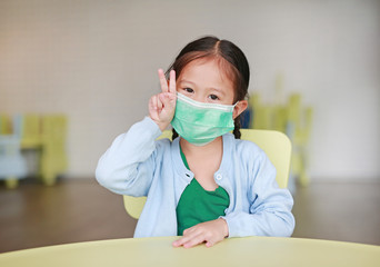 Cute little Asian child girl wearing a protective mask with showing two fingers for fight sitting on kid chair in children room.