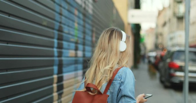 Back view on the young blonde woman in the casual clothes and headphones with a big backpack walking the street and listening to the music on the smartphone. Rear.