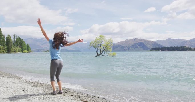New Zealand travel happy tourist woman jumping of joy at Wanaka lake landscape with lone tree, Wanaka tree famous landmark and tourist attraction.