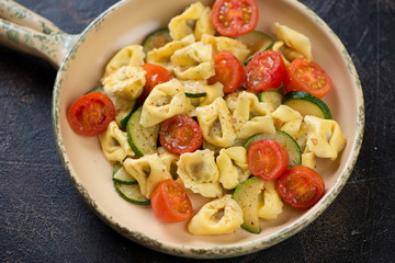 Serving pan with italian tortellini, fried zucchini and cherry tomatoes over dark brown stone surface