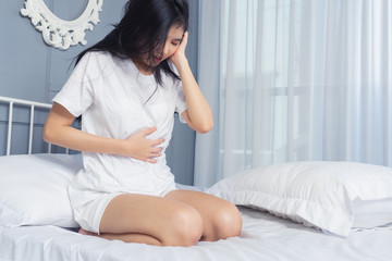 Young woman suffering from abdominal pain while sitting on white  bed at home. Do not oversleep again