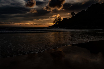 An amazing golden sunset behind a mountain silhouette edge on the end of Karon Beach, Phuket Thailand. A dramatic sky with vibrant light and reflections in the ocean