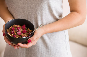 Closeup of hands of a young woman holding a black bowl with organic granola with coconut milk and raspberries. Healthy lifestyle.