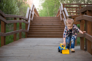 Cute boy is playing with a blue-and-yellow tractor in the park against the wooden stairs background.