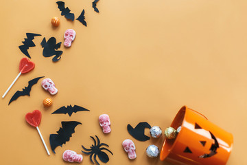 Halloween flat lay. Jack o Lantern bucket with holiday spilling  candy, bats,spiders, skulls on orange paper. Space for text. Halloween background. Season's greeting card mockup
