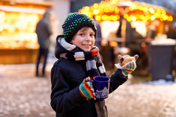 Little cute kid boy eating German sausage and drinking hot children punch on Christmas market....