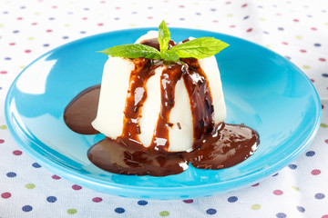 Cream pudding with chocolate sauce and mint on  multicolored tablecloth.