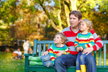Two little kid boys and young father sitting together in colorful clothing on bench. Cute healthy children, siblings and their dad having fun in autumn park on warm sunny day. Happy family of three.