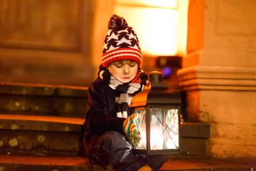 Fototapeta na wymiar Little cute kid boy with with a light lantern on stairs near church. Happy child on Christmas market in Germany. Kid waiting on parents on cold winter day..