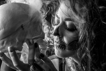 Portrait of the woman with halloween makeup close up. Talking with skull, which is in front of her. Isolated image on black background with the smoke and beautiful studio light.