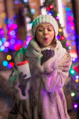 young woman in winter clothes with coffee blows snow from her hands on  background of New Year's illuminations