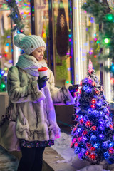 winter evening walk of  young woman with coffee near Christmas tree with  garland