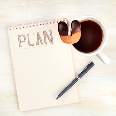 An overhead photo of a spiral notebook with the word Plan, with a cup of coffee with a cookie, shot from the top on a desk with copy space